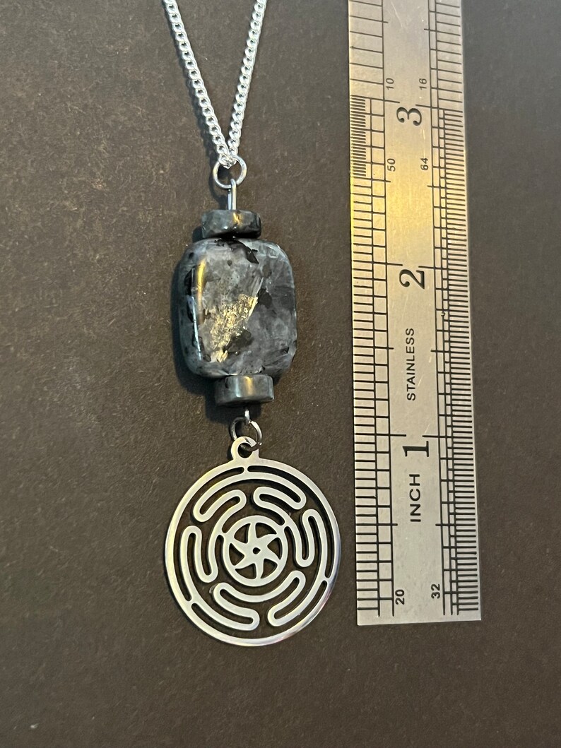 Puede incluir: Un collar de cadena de plata con un colgante de piedra negra y gris y un colgante de plata con un dise&ntilde;o circular. El collar se mide con una regla.