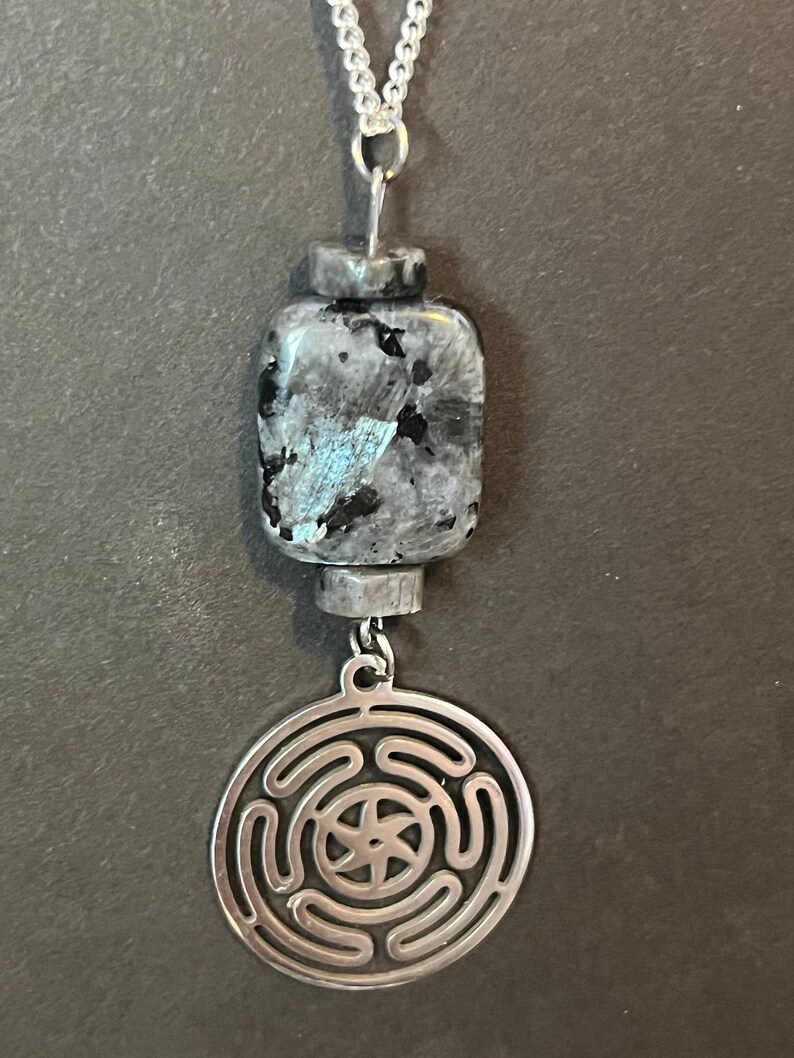 Puede incluir: Un collar de cadena de plata con un colgante de piedra gris oscuro y azul y un dije de plata con un dise&ntilde;o circular.