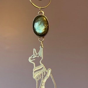 Puede incluir: Adorno dorado con un recorte de un gato egipcio con un colgante de piedra preciosa. La piedra preciosa es una labradorita verde y azul.