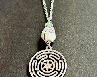 May include: Silver-toned necklace featuring a pendant with a maze-like design and a natural stone. The pendant hangs from a delicate chain. The stone is white with hints of blue and green. The necklace is displayed against a dark background.
