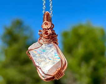 May include: A handcrafted pendant necklace featuring a clear, iridescent crystal wrapped in copper wire. The crystal is rectangular with a shimmering, rainbow effect. The pendant hangs from a delicate silver chain, perfect for adding a touch of natural beauty.