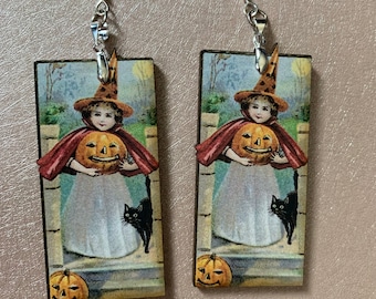 May include: A pair of Halloween-themed earrings. Each earring features a rectangular design with a vintage illustration of a child in a witch hat holding a carved pumpkin, with a black cat and a pumpkin at the bottom. The earrings have silver-colored fish hook ear wires.