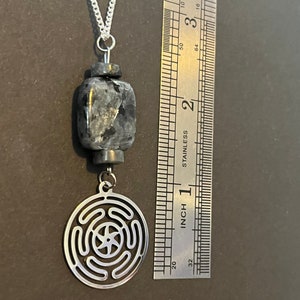 Puede incluir: Un collar de plata con un colgante de piedra negra y gris y un dije de plata con un dise&ntilde;o circular. El dije tiene una estrella de cinco puntas en el centro y c&iacute;rculos conc&eacute;ntricos alrededor.