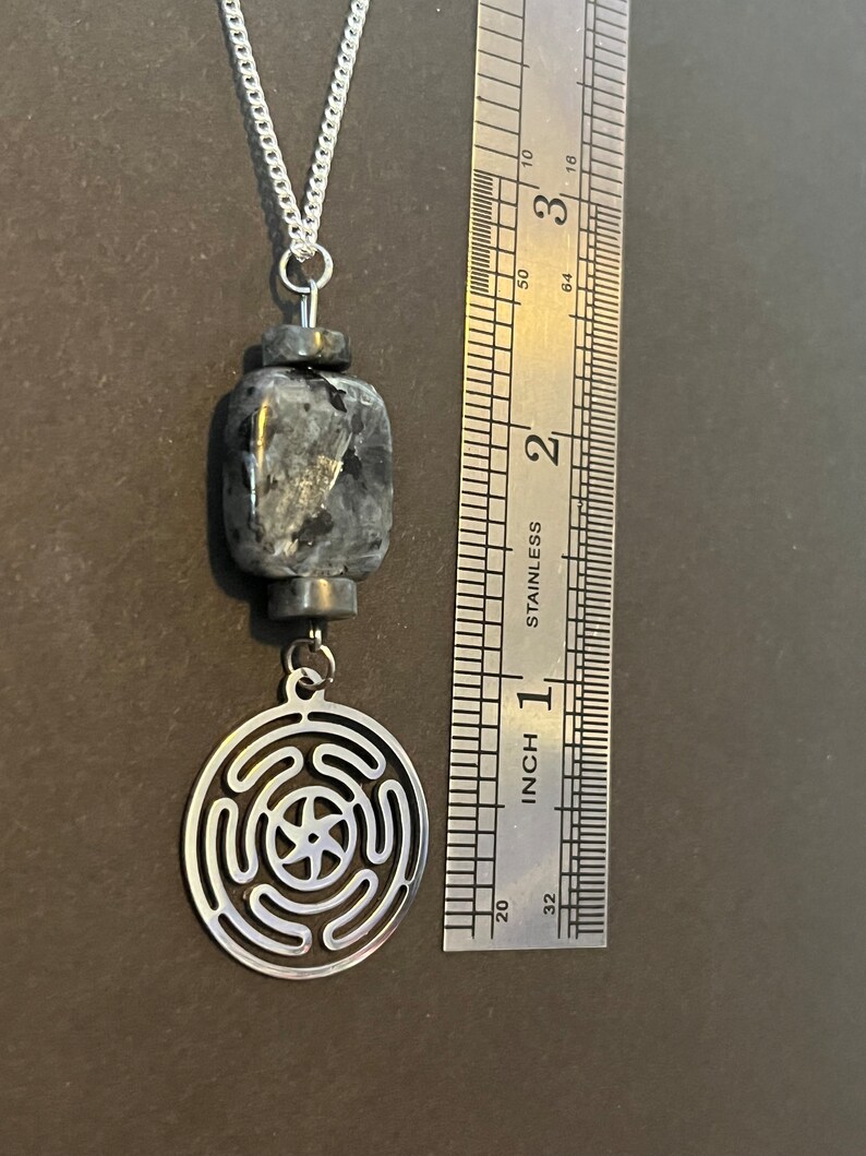 Puede incluir: Un collar de plata con un colgante de piedra negra y gris y un dije de plata con un dise&ntilde;o circular. El dije tiene una estrella de cinco puntas en el centro y c&iacute;rculos conc&eacute;ntricos alrededor.