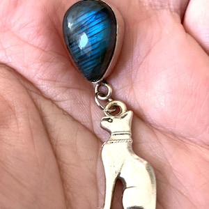 Puede incluir: Un collar de cadena de plata con un colgante en forma de lágrima con una piedra de labradorita azul. El colgante está unido a un dije de plata de un gato sentado.