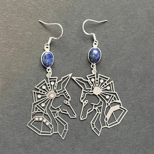 Puede incluir: Un par de pendientes de plata con un diseño geométrico que presenta una cabeza de Anubis y una piedra de lapislázuli azul.