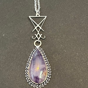Puede incluir: Collar con colgante plateado con una piedra preciosa morada en forma de lágrima. El colgante tiene un borde plateado decorativo y un símbolo estilizado. La cadena plateada fina contrasta con el fondo oscuro.