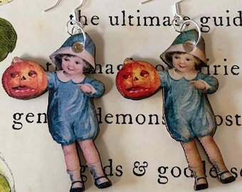 Pode incluir: Um par de brincos com tema de Halloween. Cada brinco apresenta uma imagem vintage de uma criança com roupa e chapéu azuis, segurando uma abóbora laranja esculpida. Os brincos têm fios de orelha de gancho prateados.