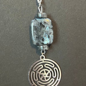 Puede incluir: Un collar de cadena de plata con un colgante de piedra azul oscuro y gris y un dije de plata con un dise&ntilde;o circular. El dije tiene una estrella de cinco puntas en el centro y cinco c&iacute;rculos conc&eacute;ntricos alrededor.