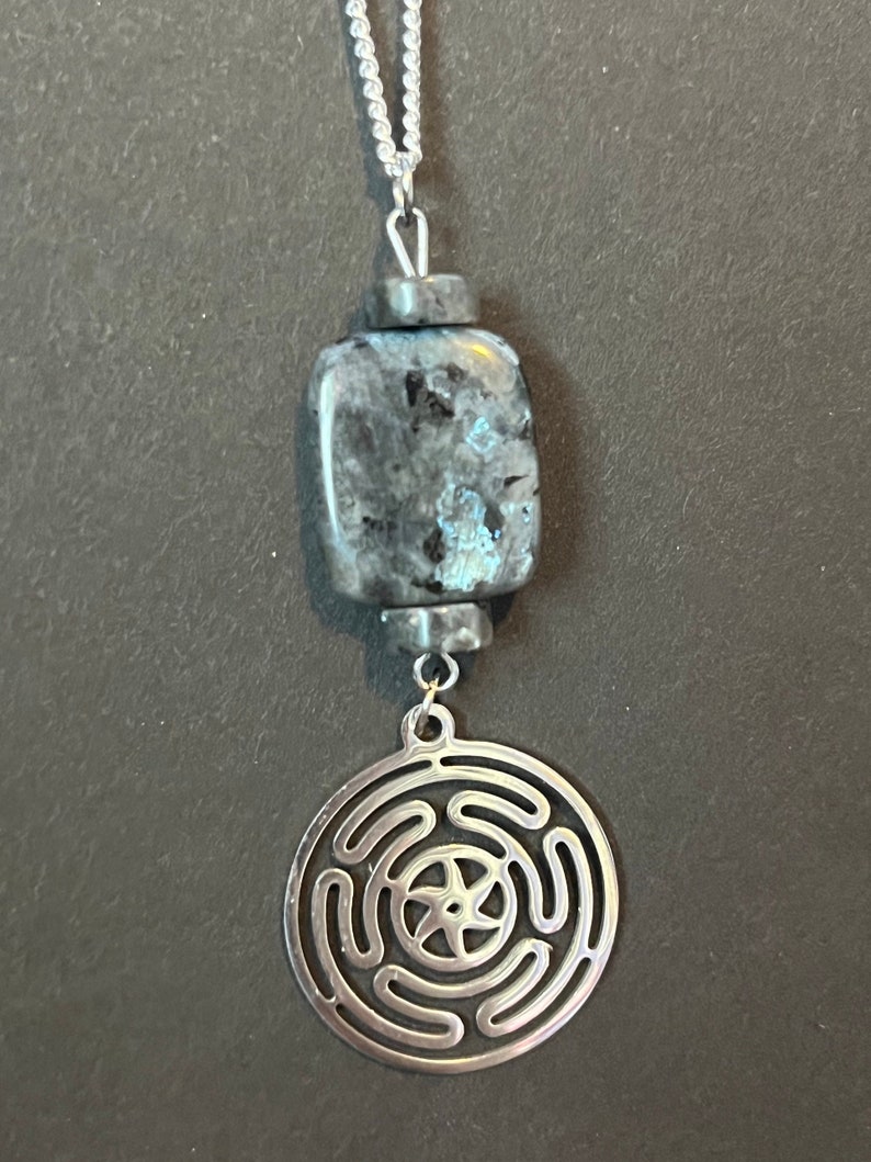 Puede incluir: Un collar de cadena de plata con un colgante de piedra azul oscuro y gris y un dije de plata con un dise&ntilde;o circular. El dije tiene una estrella de cinco puntas en el centro y cinco c&iacute;rculos conc&eacute;ntricos alrededor.