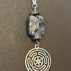 Puede incluir: Un collar de cadena de plata con un colgante de piedra negra y gris y un dije de plata con un dise&ntilde;o circular. El dije tiene una estrella de cinco puntas en el centro y cuatro c&iacute;rculos conc&eacute;ntricos alrededor.