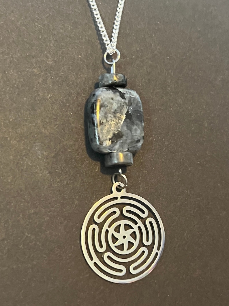 Puede incluir: Un collar de cadena de plata con un colgante de piedra negra y gris y un dije de plata con un dise&ntilde;o circular. El dije tiene una estrella de cinco puntas en el centro y cuatro c&iacute;rculos conc&eacute;ntricos alrededor.