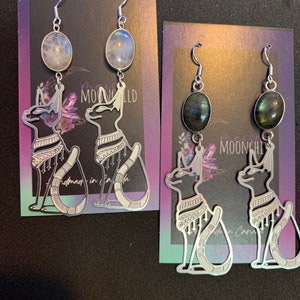 Puede incluir: Dos pares de pendientes de plata con un diseño de gato. Un par presenta una piedra de luna, el otro par presenta una piedra de labradorita. Los pendientes se muestran sobre un fondo morado y azul con el texto "MOONCHILD" visible.
