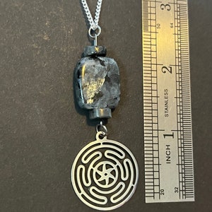 Puede incluir: Un collar de cadena de plata con un colgante negro y plateado. El colgante presenta un dije redondo de plata con un dise&ntilde;o geom&eacute;trico y una piedra cuadrada negra.