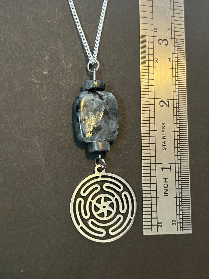 Puede incluir: Un collar de cadena de plata con un colgante negro y plateado. El colgante presenta un dije redondo de plata con un dise&ntilde;o geom&eacute;trico y una piedra cuadrada negra.