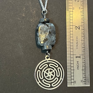 Puede incluir: Un collar de cadena de plata con un colgante negro y plateado. El colgante presenta un dise&ntilde;o circular con una estrella de cinco puntas en el centro. El colgante tiene un di&aacute;metro de aproximadamente 2,5 cm.