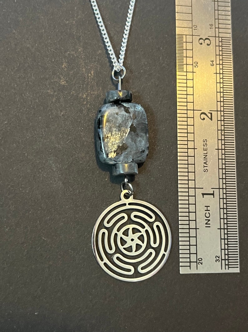 Puede incluir: Un collar de cadena de plata con un colgante negro y plateado. El colgante presenta un dise&ntilde;o circular con una estrella de cinco puntas en el centro. El colgante tiene un di&aacute;metro de aproximadamente 2,5 cm.