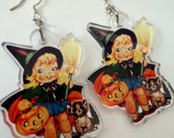 Pode incluir: Um par de brincos temáticos de Halloween com uma bruxa segurando uma abóbora e um gato. A bruxa está usando um chapéu e um vestido pretos, e os brincos são feitos de acrílico transparente.