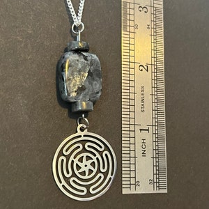 Puede incluir: Un collar de cadena plateada con un colgante negro y plateado. El colgante presenta un dise&ntilde;o circular con una estrella de cinco puntas en el centro y una piedra de forma cuadrada.