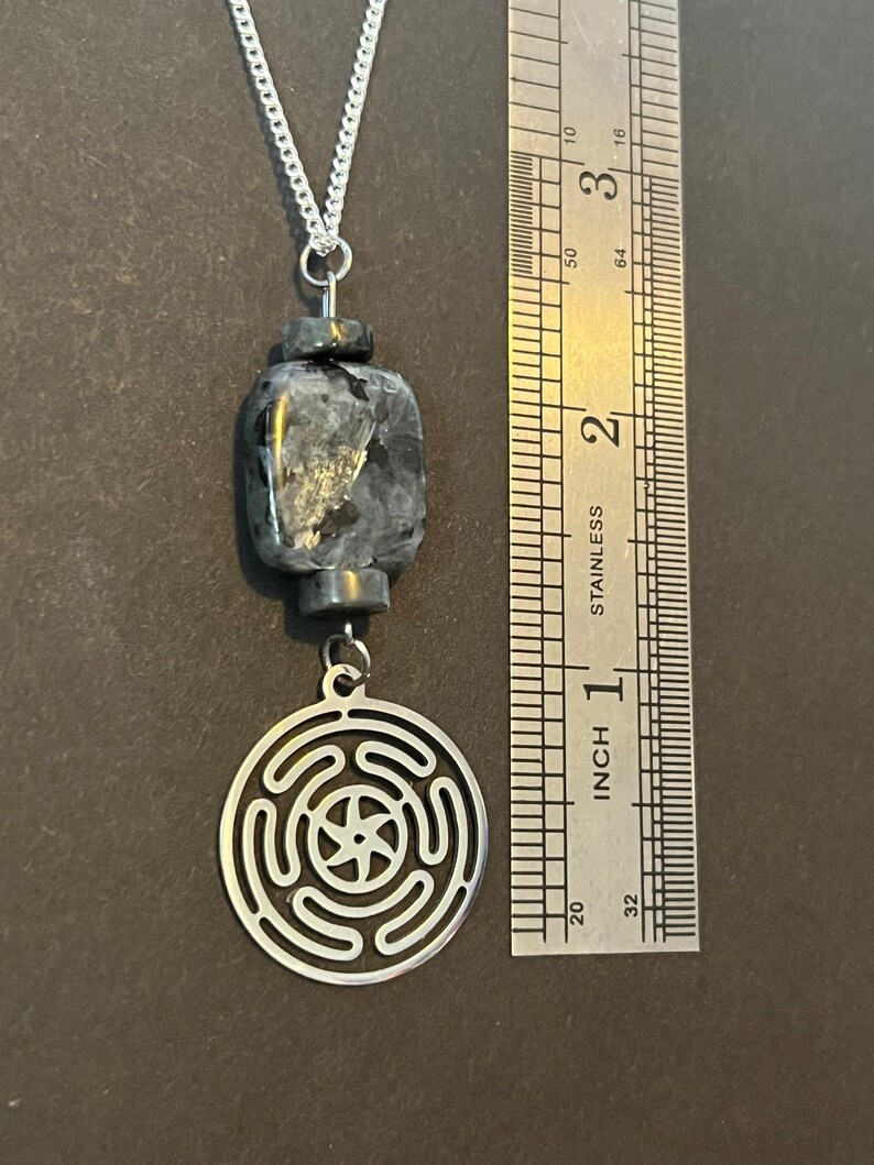 Puede incluir: Un collar de cadena plateada con un colgante negro y plateado. El colgante presenta un dise&ntilde;o circular con una estrella de cinco puntas en el centro y una piedra de forma cuadrada.