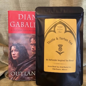 May include: A book titled "Outlander" by Diana Gabaldon, next to a black pouch of "Thistle & Tartan Tea." The tea blend is "An Outlander Inspired Tea Blend" with Assam Black Tea, dried blueberries, elderflowers, and hibiscus.