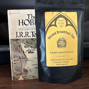 May include: A copy of The Hobbit by J.R.R. Tolkien next to a black pouch of Second Breakfast Tea. The yellow label features a round door illustration and text listing ingredients including English Breakfast Tea, Red Rooibus, Cinnamon, Dried Apple, and Dried Orange Peel.