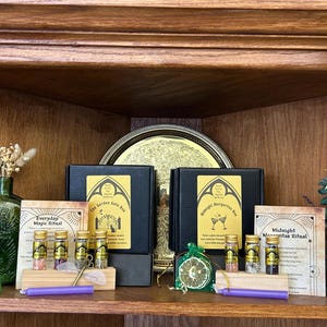 May include: Two black square boxes with gold labels titled The Garden Gate Box and Midnight Margaritas Box, accompanied by small glass vials, ritual cards, purple candles, and dried botanical elements arranged on a wooden shelf.