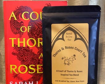 Może przedstawiać: Czerwona książka zatytułowana A Court of Thorns and Roses autorstwa Sarah J. Maas obok czarnej torebki herbaty Thorns & Roses Court Tea. Etykieta zawiera gotycki łuk, tekst Rooted In Love Teas oraz składniki Irish Breakfast Tea, Cloves, Rose Petals.