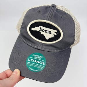 May include: Gray and white trucker hat with a black oval patch featuring the outline of North Carolina and the text "home."