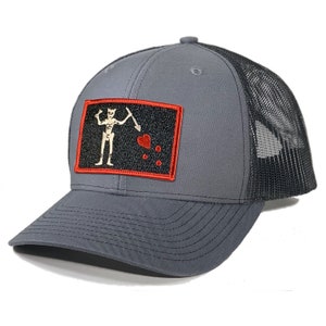 May include: A gray and black trucker hat with a red-bordered patch featuring a black and white skeleton holding a sword and a red heart. The patch has a black background with white dots.