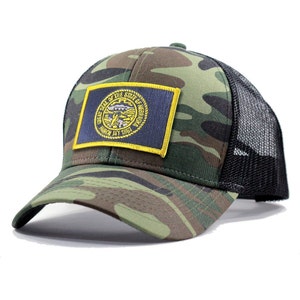 May include: A camouflage trucker hat with a black mesh back. The hat features a patch with the Great Seal of the State of Nebraska.