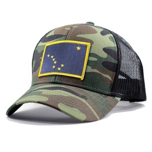 Peut inclure: Un chapeau de camionneur camouflage avec un dos en maille noire et un patch jaune représentant le drapeau de l'État d'Alaska. Le drapeau est bleu avec une étoile dorée et huit étoiles dorées en forme de la constellation de la Grande Ourse.