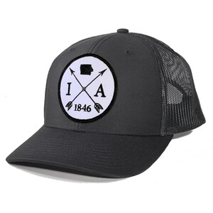 May include: A dark gray trucker hat with a white patch featuring a black outline of the state of Iowa, crossed arrows, the letters "IA", and the year "1846".