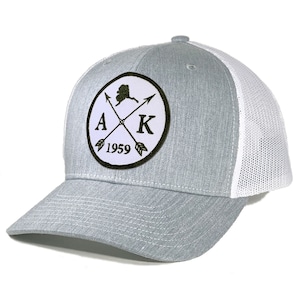 May include: A grey and white trucker hat with a white patch featuring a black outline of Alaska, the letters "AK", two crossed arrows, and the year "1959".