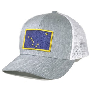 Peut inclure: Un chapeau de camionneur gris et blanc avec un écusson brodé bleu et or du drapeau de l'État d'Alaska. Le drapeau présente un fond bleu avec un contour doré et huit étoiles dorées représentant la constellation de la Grande Ourse.