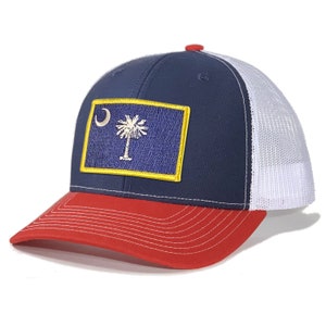 May include: A navy blue and red trucker hat with a white mesh back. The hat features a yellow-trimmed patch with the South Carolina state flag.