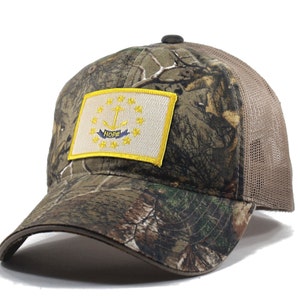 May include: A camouflage trucker hat with a brown mesh back. The front of the hat features a yellow embroidered patch of the Rhode Island state flag with the words "Hope" on the anchor.