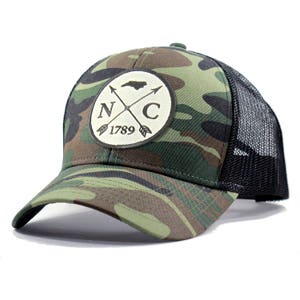 May include: A camouflage trucker hat with a black mesh back. The front features a white patch with a black outline and the text "NC 1789" and a silhouette of the state of North Carolina with two crossed arrows.