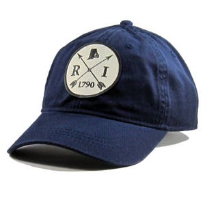 May include: A navy blue baseball cap with a white patch featuring a crossed arrow design, the letters "RI", and the year "1790". The patch also includes an outline of the state of Rhode Island.