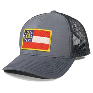 May include: A gray and black trucker hat with a patch of the Georgia state flag on the front. The flag is red, white, and blue with a gold emblem in the center.