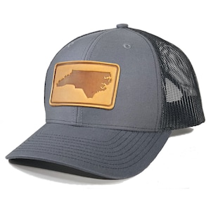 May include: A grey and black trucker hat with a brown leather patch featuring the outline of North Carolina.