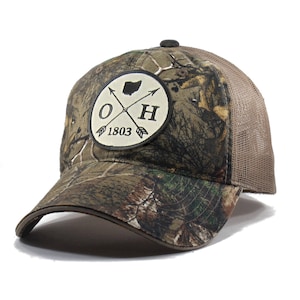 May include: A brown and green camouflage trucker hat with a beige mesh back. The hat features a white patch with a black outline and the text "OH 1803" and a crossed arrow design.