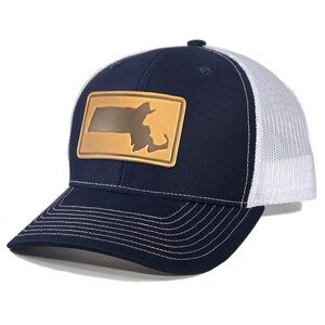 May include: A navy blue and white trucker hat with a gold-colored patch featuring the outline of Massachusetts. The hat has a curved brim with white stitching and a white mesh back.