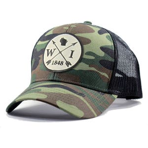 May include: A camouflage trucker hat with a black mesh back. The front of the hat features a circular patch with a white background and black text that reads "WI 1848". Inside the circle is a black outline of the state of Wisconsin and two crossed arrows.