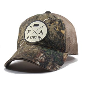 May include: A brown and green camouflage trucker hat with a white patch featuring a black outline of the state of Pennsylvania, two crossed arrows, and the text "PA 1787".