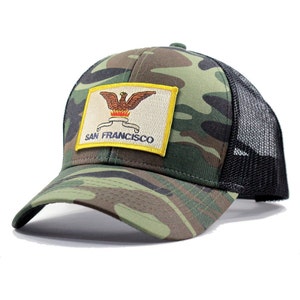 May include: A camouflage trucker hat with a black mesh back. The hat features a yellow patch with the San Francisco city seal and the text "SAN FRANCISCO".