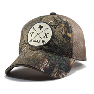 May include: A brown and green camouflage trucker hat with a beige mesh back. The hat features a round patch with a white background and black text that reads "TX 1845" with a stylized Texas outline and crossed arrows.