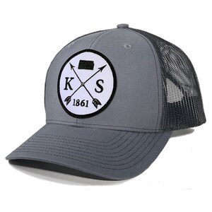 May include: A gray and black trucker hat with a white patch featuring a black outline of the state of Kansas, crossed arrows, the letters "K" and "S", and the year "1861".