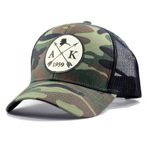 May include: A camouflage trucker hat with a black mesh back. The front of the hat features a white circle with a black outline and the text "AK 1959" inside. A black silhouette of Alaska with two crossed arrows is also inside the circle.