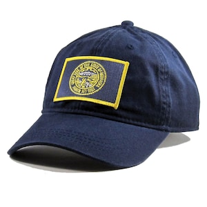 May include: A navy blue baseball cap with a yellow embroidered patch featuring the Great Seal of the State of Nebraska. The seal depicts a  bunting, a plow, a sheaf of wheat, and a river. The text on the seal reads "Great Seal of the State of Nebraska March 1st 1867".